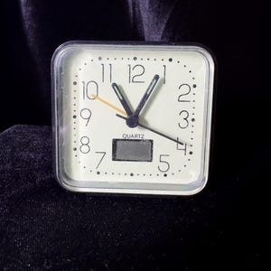 May include: A square quartz alarm clock with a cream-colored face, black numerals, and a small digital display. The clock has a clear plastic frame and black hands, with a yellow second hand. The clock is approximately 3 inches wide.