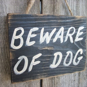 May include: A black wooden sign with white painted text that reads "Beware of Dog". The sign is hanging from a rope.