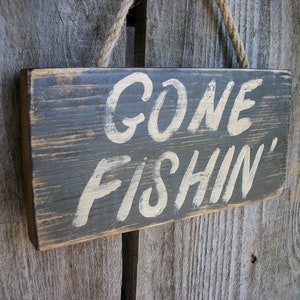 May include: A rustic wooden sign with a distressed gray paint finish. The sign has white lettering that reads "Gone Fishin'" and is hanging from a piece of twine.