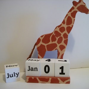 May include: A wooden giraffe figurine with brown and tan spots stands on a white block calendar with the months of January, May, July, and November displayed. The calendar also shows the date as 4-01.