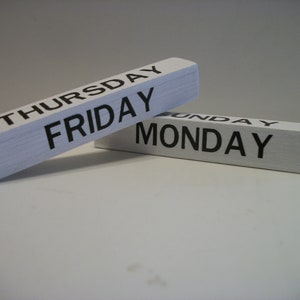 May include: Two white wooden blocks with black lettering. The top block reads "THURSDAY FRIDAY" and the bottom block reads "SUNDAY MONDAY".
