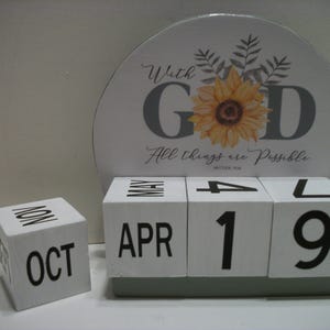 May include: A white wooden block calendar displaying "APR 19" with a separate block showing "OCT NOV". A decorative sign behind the calendar features a sunflower and the words "With GOD All things are Possible".