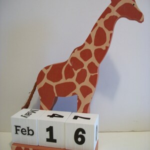 May include: A wooden giraffe figurine with a brown and tan patterned design holds a white block calendar with the date February 16th.