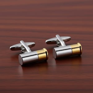 Może przedstawiać: Para srebrnych i złotych spinek do mankietów. Spinki do mankietów mają kształt pocisku ze srebrnym korpusem i złotymi akcentami. Spinki do mankietów leżą na ciemnobrązowej drewnianej powierzchni.