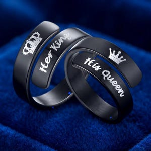 May include: Two black rings with a matte finish. One ring features a white engraved crown and the text Her King. The other ring features a white engraved crown and the text His Queen. Both rings have a bypass design with overlapping ends.