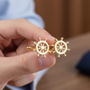Può includere: Un paio di gemelli dorati a forma di timoni di nave. I gemelli sono tenuti in mano, mostrando il loro design intricato e la finitura lucida. Questi accessori a tema nautico sono un'aggiunta classica ed elegante a qualsiasi abito formale.