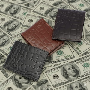 Puede incluir: Tres carteras de cuero en tonos negro, marrón y marrón oscuro, cada una con una textura de piel de cocodrilo. Las carteras se exhiben sobre un fondo de billetes de cien dólares dispersos.