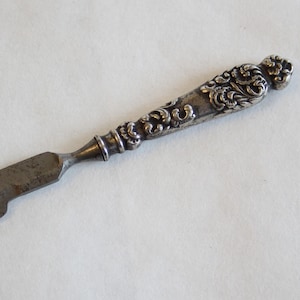 May include: A silver-toned, ornate, decorative knife with a pointed blade and a detailed, floral handle.