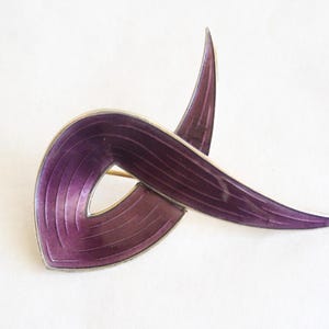 Peut inclure: Broche vintage en émail violet avec une bordure en métal argenté. La broche présente un design abstrait avec des lignes courbes et fluides. L'émail a un aspect texturé, et la broche est posée sur un fond blanc.