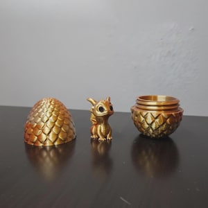 May include: A golden dragon figurine, a dragon egg, and a container with a dragon egg design are displayed. The dragon has large eyes and detailed wings. The egg and container have a textured, scale-like pattern.