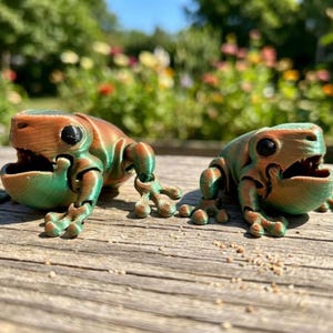 May include: Two articulated frog figurines featuring a dual-tone color scheme of metallic copper and emerald green. Each frog has large black eyes, a wide open mouth, and visible horizontal layer lines from the 3D printing process.