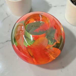 Op de afbeelding: Een ronde kandelaar met een levendig ontwerp. De houder is gemaakt van heldere hars, met ingebedde oranje en groene bloemelementen. De kandelaar heeft een diameter van ongeveer 7,6 cm.