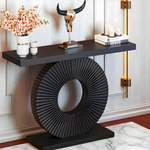 May include: A black console table with a unique circular base design. The table features a dark gray top with decorative items, including a gold bull skull sculpture, vases with flowers, and candles. Two gold wall sconces are mounted on the wall.