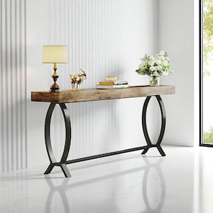 May include: A wooden console table with a dark metal base. The tabletop is made of light brown wood, and the base has a unique, curved design. A lamp, decorative figurine, books, and flowers adorn the table, creating a stylish display.