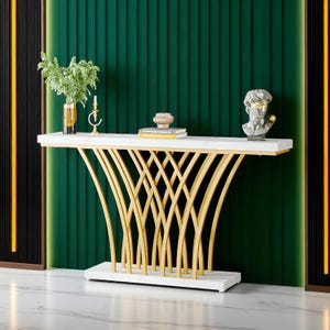 Puede incluir: Una consola con tapa de mármol blanco y base dorada entrelazada. La mesa está decorada con un jarrón con vegetación, un candelabro, una pequeña escultura y libros. El fondo es una pared verde oscuro.