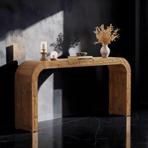May include: A curved wooden console table with a natural grain finish. On the surface sits a tall candle holder, a small glass candle votive, a closed book, a small bowl, and a white ceramic vase containing a dried floral arrangement.
