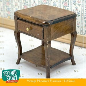 May include: A miniature vintage wooden nightstand with a drawer and lower shelf. The nightstand is brown with a dark trim and a gold-colored knob. The legs are curved. The text "Second Story miniatures" and "Vintage Miniature Furniture - 1:12 Scale" are visible.