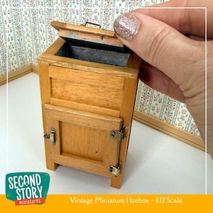 Peut inclure: Un petit réfrigérateur en bois avec un couvercle et une porte à charnières, avec une quincaillerie en métal. Le réfrigérateur est brun clair avec un intérieur sombre. Le texte "Vintage Miniature | Icebox - 1:12 Scale" est visible.