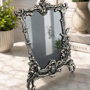 May include: A rectangular tabletop mirror featuring an ornate, silver-toned frame with intricate relief carvings, scrollwork, and floral motifs. The mirror stands on two decorative feet and has a reflective glass surface with a dark, smooth finish.