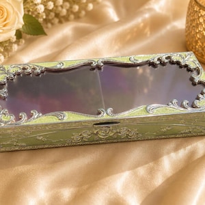 May include: A rectangular storage box featuring a light green enamel finish with intricate silver-toned metal filigree detailing. The lid includes a clear, dark-tinted glass panel framed by ornate scrollwork and embossed floral patterns.