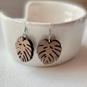May include: A pair of wooden earrings shaped like monstera leaves. The earrings are brown and have a silver hook.