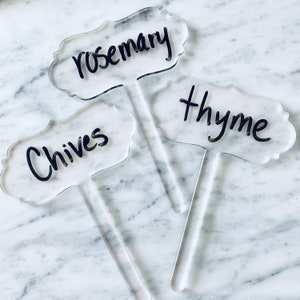 May include: Three clear acrylic plant markers with black handwritten labels. The labels read "rosemary", "thyme", and "chives".