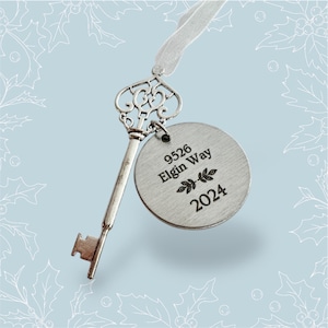 May include: A silver key ornament with a round tag attached. The tag is engraved with the text "9526 Elgin Way 2024" and a small leaf design.