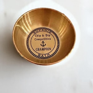 May include: A small, round, gold-colored bowl with a slightly flared rim. The bowl's interior features a circular design with the words "SUPSHIP Chip & Dip Competition CHAMPION BATH" and an anchor symbol. The bowl is likely made of metal.