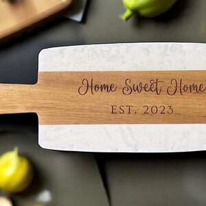 May include: A rectangular wooden cutting board with a handle. The board features a white marble-like surface on the top and bottom, with a wooden stripe in the middle. The text "Home Sweet Home EST. 2023" is engraved on the wooden stripe.