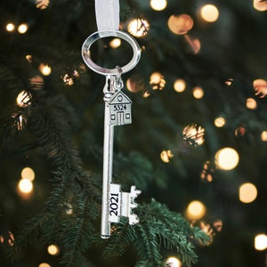 May include: Silver key-shaped Christmas ornament with a house charm and the year 2021 engraved on the key. The ornament is hanging from a Christmas tree with twinkling lights.