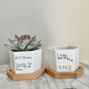 May include: Two white hexagonal ceramic planters with handwritten messages. The planter on the left has a succulent plant and reads "MRS. BRUANT Thanks for Helping Me Grow". The planter on the right reads "Love Matthew 2023". Both planters are on wooden hexagonal coasters.