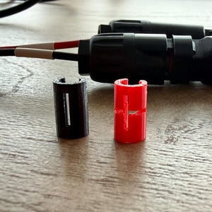 以下が含まれることがあります： 電気コネクタとコンポーネントのクローズアップ。画像には、黒いコネクタ、2つの小さな円筒形の部品（1つは黒、1つは赤）、赤と白の絶縁を備えたワイヤーが写っています。コンポーネントは木製の表面にあります。