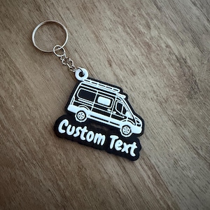 May include: A black and white van-shaped keyring with the text "Custom Text". The keyring has a silver key ring and chain. The van design includes wheels, windows, and a roof rack. The keyring is on a wooden surface.