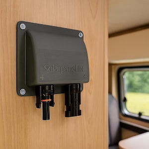 May include: A dark gray solar panel IN junction box mounted on a light wood surface. The box has two black connectors and the text "Solarpanel IN". The box is secured with four screws.