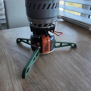 May include: A compact camping stove with a grey burner and a green base. The stove is mounted on a black and orange fuel canister. A red wire extends from the burner. The stove is on a wooden surface.