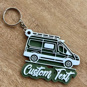 May include: A green and white camper van keychain with the words "Custom Text" in a cursive font. The keychain has a silver ring and chain. The van has a detailed design with wheels, windows, and a roof rack.