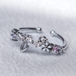 Puede incluir: Anillo ajustable plateado con diseño floral. El anillo está adornado con piedras preciosas claras y rosas, junto con flores de esmalte blanco. El anillo tiene una estética delicada y femenina.