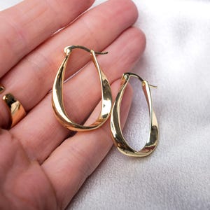 Könnte beinhalten: Ein Paar goldfarbene, gedrehte ovale Creolen. Die Ohrringe haben eine polierte Oberfläche und ein schlichtes, elegantes Design. Die Ohrringe werden vor einem weichen, hellen Hintergrund gezeigt.