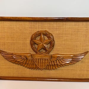 May include: Wooden wall art featuring a carved emblem with a star, laurel wreath, and wings. The emblem is set against a tan fabric background within a brown wooden frame. The artwork is a decorative piece.