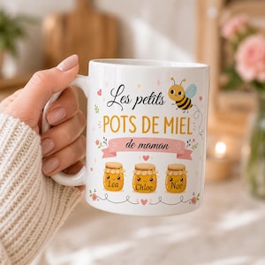 May include: White ceramic mug with the text "Les petits pots de miel de maman" and three honey jar illustrations with the names Lea, Chloe, and Noe. A cartoon bee is also featured on the mug. The mug is held by a person.