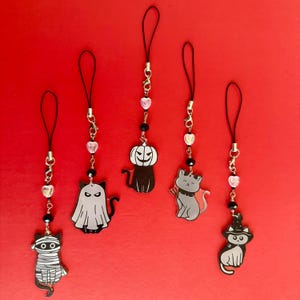 May include: Five phone charms featuring cat characters in Halloween costumes, including a mummy, a ghost, a pumpkin-headed cat, a devil cat, and a witch cat. Each charm hangs from a black cord with gold-tone hardware, a heart bead, and a black bead.