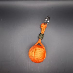 Op de afbeelding: Een oranje paracord monkey fist sleutelhanger met een zilveren sleutelring. De knoop is geaccentueerd met een gedeelte olijfgroen koord. De monkey fist is een decoratieve knoop die als sleutelhanger wordt gebruikt.