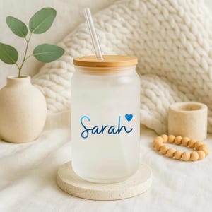 May include: A frosted glass tumbler with a bamboo lid and a clear glass straw. The tumbler features the name Sarah in a blue cursive script followed by a small blue heart. The glass has a smooth, matte finish and a cylindrical shape.