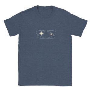 Peut inclure: Un t-shirt bleu marine avec un motif céleste. Le motif comprend deux étoiles à l'intérieur d'une forme ovale double. Les étoiles sont de couleur beige clair et les ovales sont gris clair.