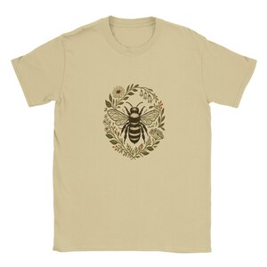 Pode incluir: Uma camiseta bege com uma ilustração detalhada de uma abelha dentro de uma coroa de flores. A abelha é retratada em preto e branco, cercada por folhas verdes e pequenas flores. A camiseta tem gola redonda clássica e mangas curtas.
