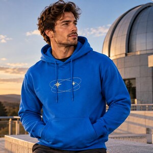 Pode incluir: Uma camisola com capuz azul brilhante com um gráfico branco de duas órbitas elípticas que se cruzam com estrelas de quatro pontas no peito. A peça inclui um bolso frontal tipo canguru e cordões azuis a condizer no decote.