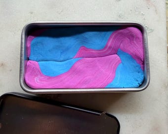 May include: A rectangular metal container filled with blue and pink kinetic sand. The lid is open and placed next to the container. The sand has a marbled appearance, with the colors swirled together. The container sits on a white surface.