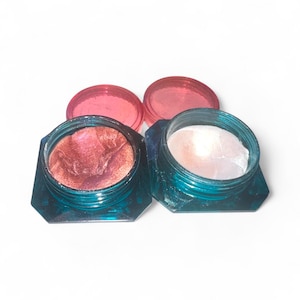 May include: Two open jars of cosmetic products with teal-colored bases and pink lids. One jar contains a shimmery, reddish-brown product, and the other has a pearlescent, white product. The jars are on a white background.