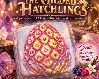 May include: A square clear container holding putty with a sculpted dragon egg design featuring gold and pink scales accented with embedded crystals. The label reads The Gilded Hatchlings, Mega Dragon Drill-A-Long, Tatertottz Creationz, and Scent: Blackberry Raspberry Vanilla.