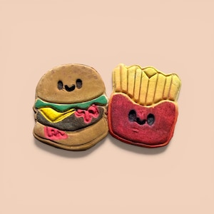 May include: Two food-themed cookies: a smiling hamburger with lettuce, cheese, and sauce, and a container of smiling french fries. The cookies are brown, yellow, green, and red, with black facial features. The background is a light peach color.